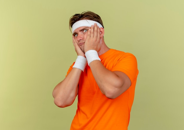 Impressed young handsome sporty man wearing headband and wristbands putting hands on cheeks isolated on olive green