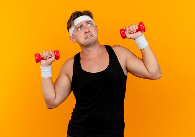 Impressed young handsome sporty man wearing headband and wristbands raising dumbbells looking at side isolated on orange