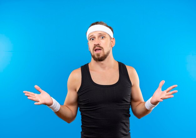 Impressed young handsome sporty man wearing headband and wristbands showing empty hands isolated on blue wall