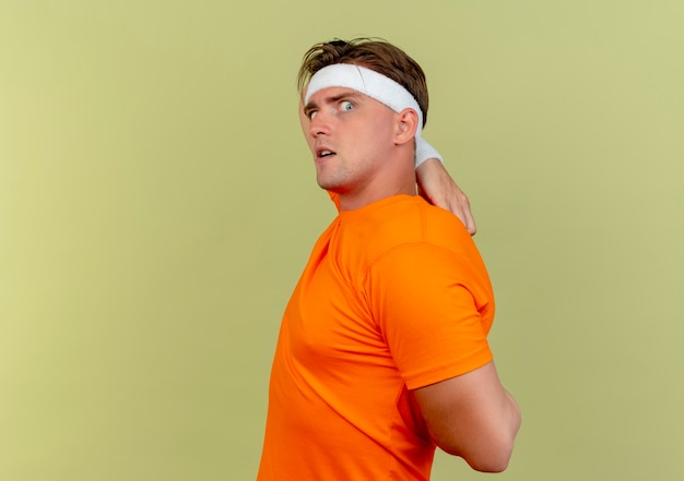 Free photo impressed young handsome sporty man wearing headband and wristbands standing in profile view putting hands on back isolated on olive green