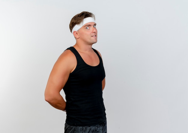 Impressed young handsome sporty man wearing headband and wristbands standing in profile view putting hands behind back isolated on white