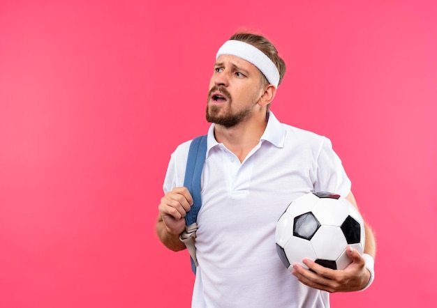 Впечатленный молодой красивый спортивный мужчина с головной повязкой и браслетами с задней сумкой на плече, держащий футбольный мяч, смотрящий в сторону, изолированную на розовой стене с копией пространства
