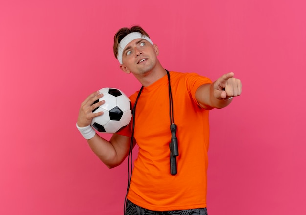 Impressed young handsome sporty man wearing headband and wristbands with jump rope around neck holding soccer ball pointing and looking at side isolated on pink