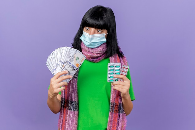 Impressed young ill caucasian girl wearing mask and scarf holding money and packs of capsules looking at money isolated on purple background with copy space