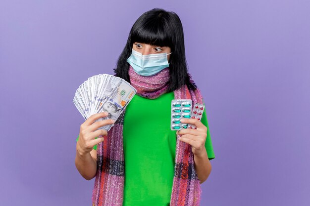 Впечатленная молодая больная кавказская девушка в маске и шарфе держит деньги и пачки капсул, глядя на деньги, изолированные на фиолетовом фоне с копией пространства
