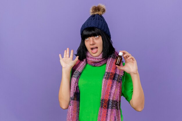 Impressed young ill caucasian girl wearing winter hat and scarf holding medicament in glass showing empty hand looking up isolated on purple background with copy space