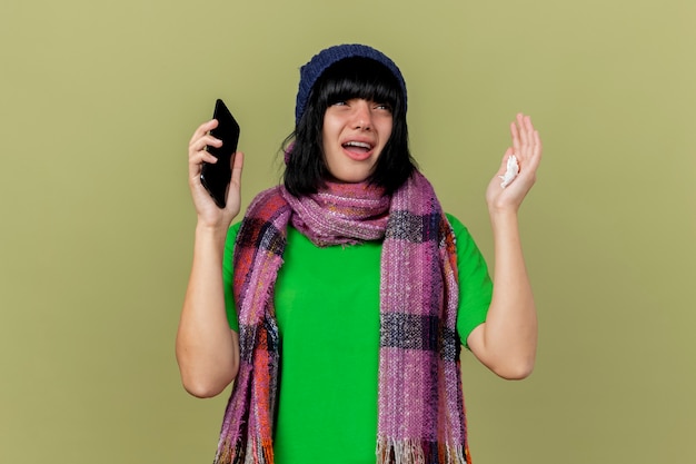 コピースペースでオリーブグリーンの背景に分離された手を上げる側を見て携帯電話とナプキンを保持している冬の帽子とスカーフを身に着けている印象的な若い病気の白人の女の子