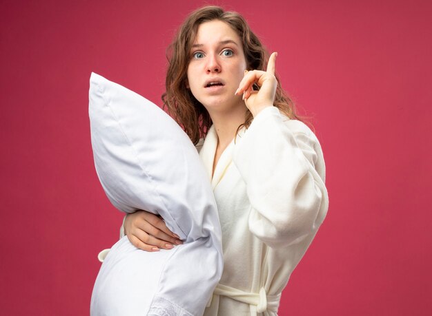 Impressed young ill girl looking straight ahead wearing white robe hugged pillow points at up isolated on pink