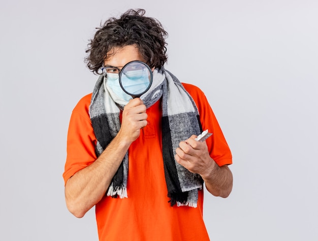 Free photo impressed young ill man wearing glasses scarf and mask holding packs of medical pills looking at them through magnifying glass isolated on white wall