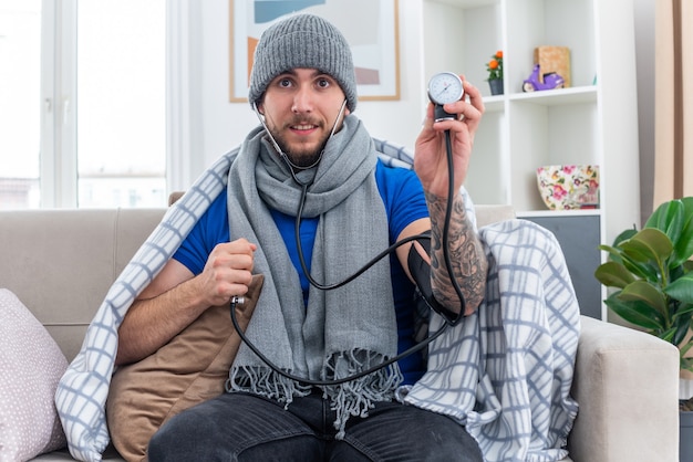 Impressed young ill man wearing scarf and winter hat with stethoscope wrapped in blanket sitting on sofa in living room showing sphygmomanometer