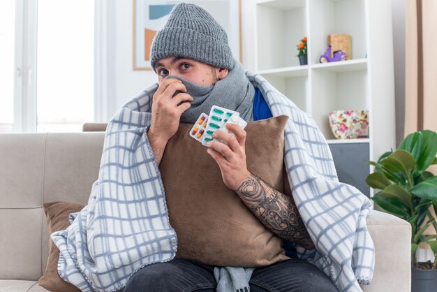 Impressed young ill man wearing scarf and winter hat wrapped in blanket sitting on sofa in living room holding pillow covering mouth and nose with scarf looking up holding packs of pills