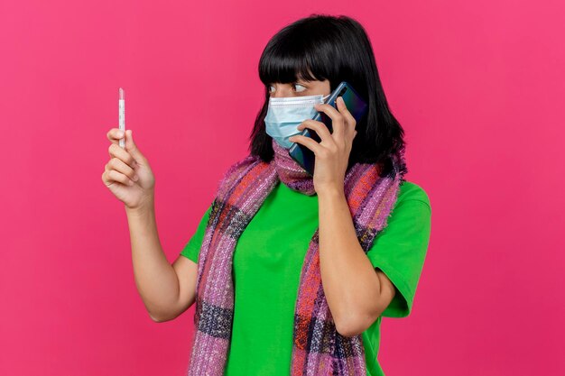 コピースペースのあるピンクの壁に隔離された電話で話している温度計を持って見ているマスクとスカーフを身に着けている感動の若い病気の女性