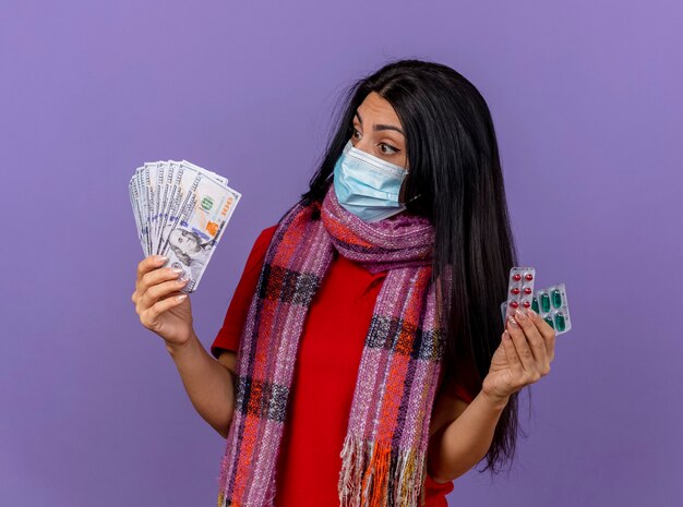 Impressed young ill woman wearing mask and scarf holding money and pack of capsules looking at money isolated on purple wall