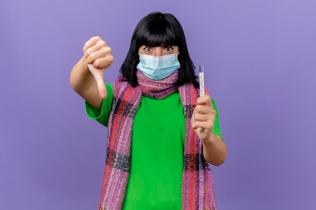 Free photo impressed young ill woman wearing mask and scarf holding thermometer looking at front showing thumb down isolated on purple wall with copy space