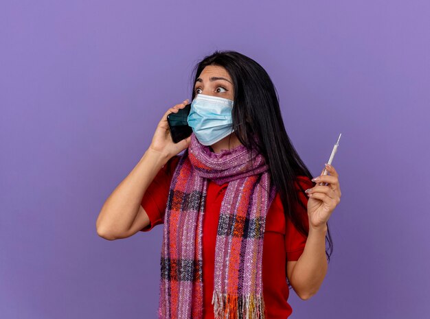 紫色の壁に隔離された側を見て電話で話している温度計を保持しているマスクとスカーフを身に着けている感銘を受けた若い病気の女性