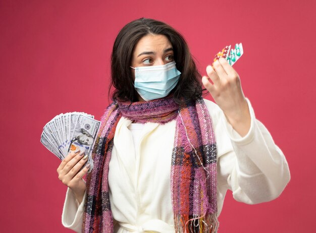 Impressed young ill woman wearing robe and scarf with mask holding money and packs of medical capsules looking at capsules isolated on pink wall