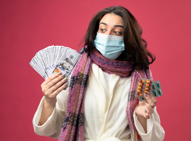 Impressionato giovane donna malata che indossa accappatoio e sciarpa con maschera che tiene soldi e confezioni di capsule mediche guardando soldi isolati sul muro rosa