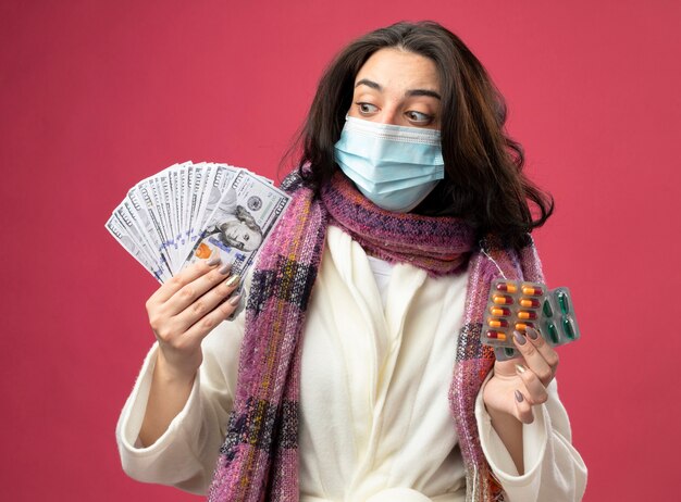 Impressed young ill woman wearing robe and scarf with mask holding money and packs of medical capsules looking at money isolated on pink wall