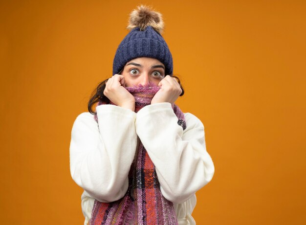 Impressed young ill woman wearing robe winter hat and scarf looking at front covering mouth with scarf isolated on orange wall