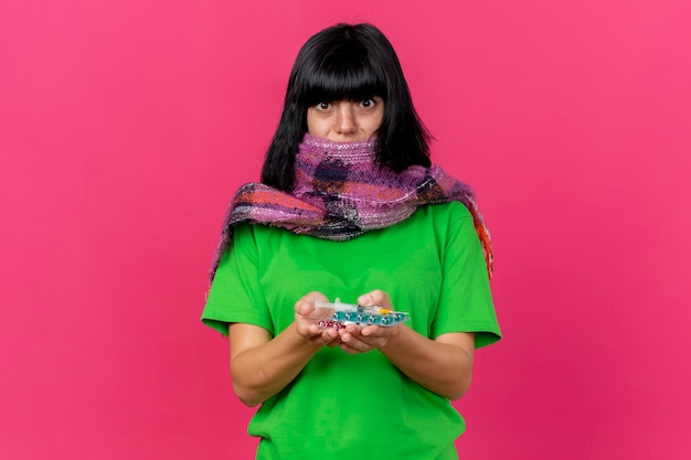 Impressed young ill woman wearing scarf holding syringe and medical pills looking at front isolated on pink wall with copy space