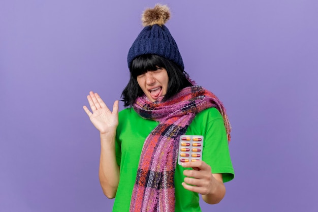 Impressed young ill woman wearing winter hat and scarf holding pack of capsules looking at front showing empty hand isolated on purple wall with copy space