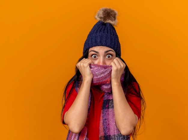 Foto gratuita impressionato giovane donna malata che indossa cappello invernale e sciarpa guardando la bocca della copertura anteriore con sciarpa isolata sulla parete arancione