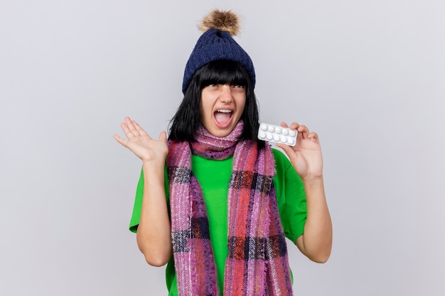 Impressed young ill woman wearing winter hat and scarf showing pack of tablets and empty hand looking up isolated on white wall