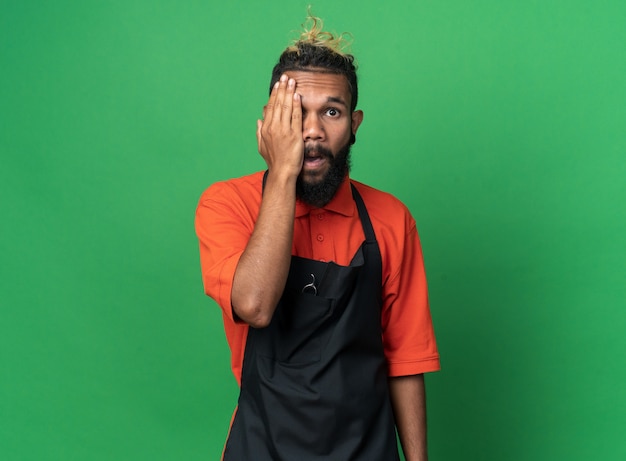 Impressed young male barber wearing uniform covering half of face with hand looking at front isolated on green wall with copy space