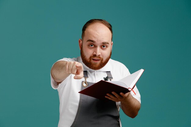 Impressed young male barber wearing white shirt and barber apron holding note pad looking and pointing at camera isolated on blue background