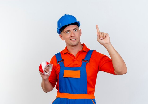 impressed young male builder wearing uniform and safety helmet holding duct tape and points at up