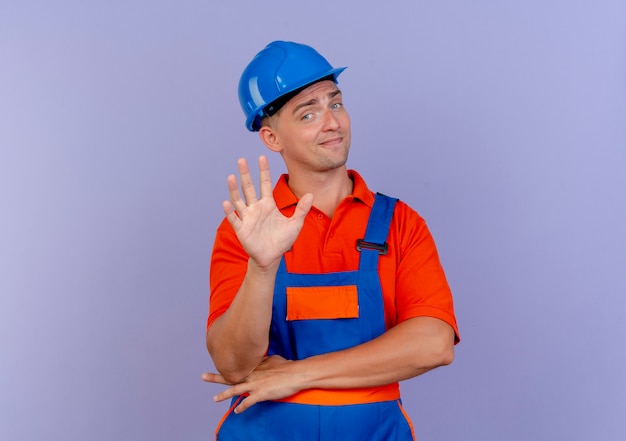 впечатленный молодой мужчина-строитель в форме и защитном шлеме, показывающий пять