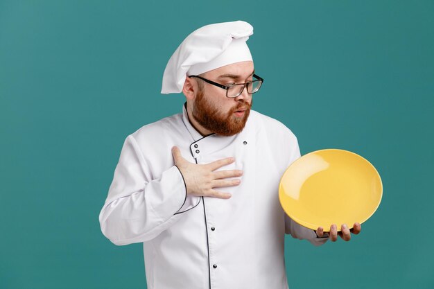 Impressionato giovane chef maschio che indossa occhiali uniformi e cappuccio che tiene piatto vuoto tenendo la mano sul petto guardando il piatto isolato su sfondo blu