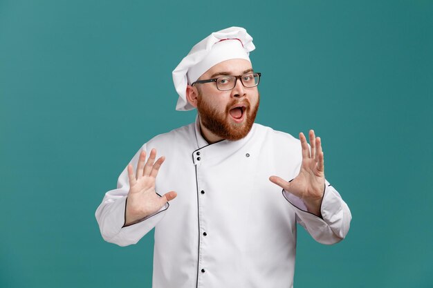 Impressed young male chef wearing glasses uniform and cap looking at camera showing empty hands isolated on blue background