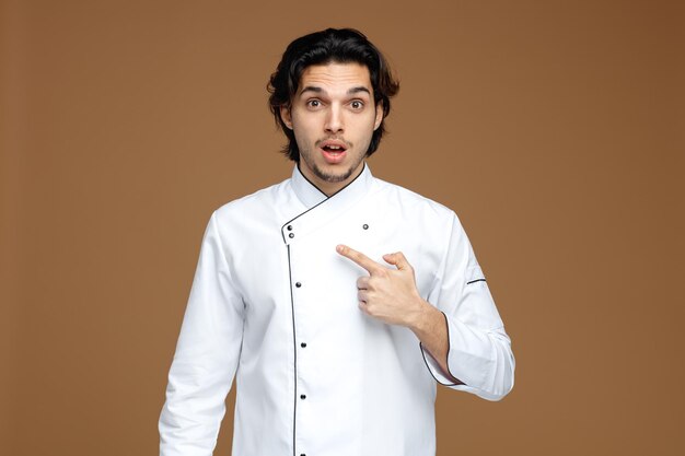 impressed young male chef wearing uniform looking at camera pointing at himself isolated on brown background