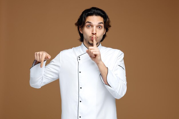 impressed young male chef wearing uniform looking at camera showing silence gesture pointing down isolated on brown background