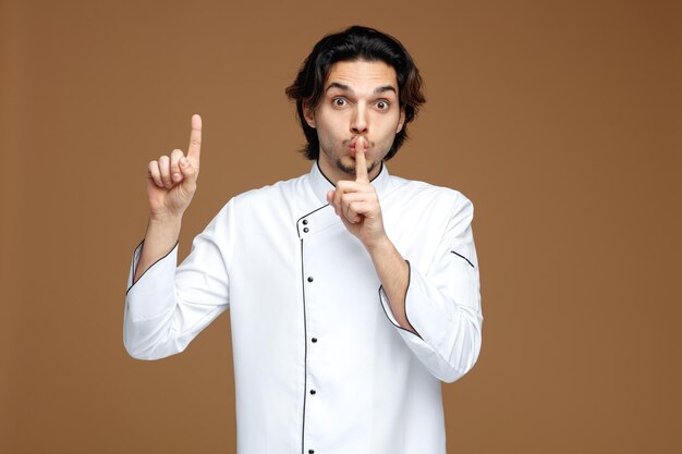 впечатленный молодой шеф-повар в униформе смотрит в камеру, показывая жест молчания, указывающий вверх на коричневом фоне