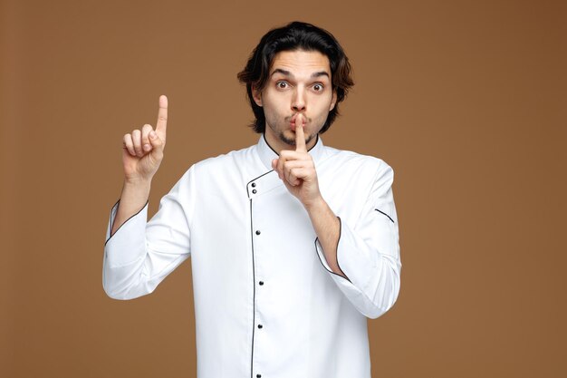 impressed young male chef wearing uniform looking at camera showing silence gesture pointing up isolated on brown background