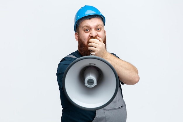 Impressed young male construction worker wearing safety helmet and uniform stretching loudspeaker out towards camera looking at camera talking into speaker isolated on white background