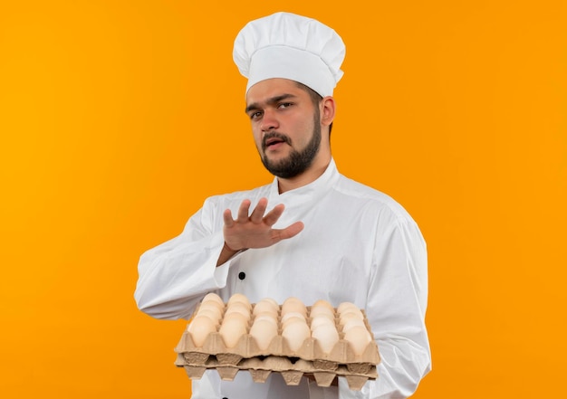 Impressionato giovane cuoco maschio in uniforme da chef che tiene una scatola di uova tenendo la mano sopra di loro isolata sulla parete arancione