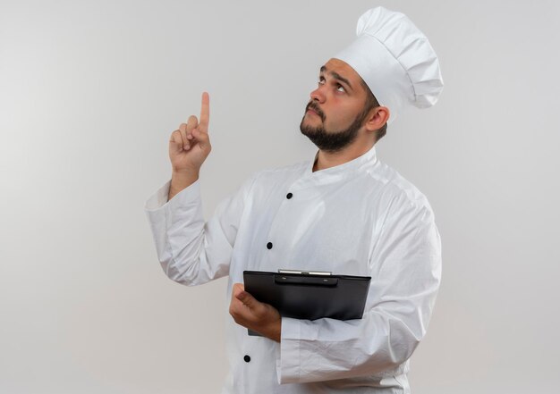 Impressed young male cook in chef uniform holding clipboard looking and pointing up isolated on white wall with copy space