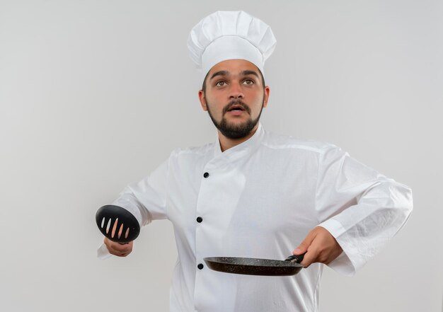 Impressed young male cook in chef uniform holding slotted spoon and frying pan looking up isolated on white wall