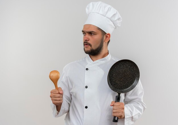 Впечатленный молодой мужчина-повар в униформе шеф-повара держит ложку и сковороду на белой стене с копией пространства
