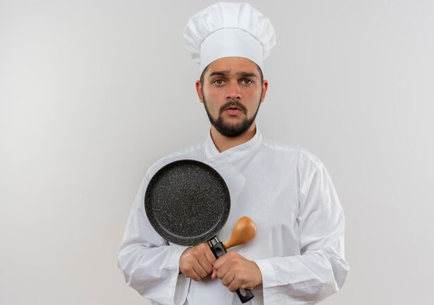 Impressed young male cook in chef uniform holding spoon and frying pan isolated on white wall with copy space