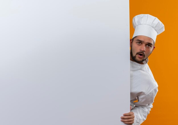 Impressed young male cook in chef uniform looking from behind and putting hand on white wall isolated on orange wall with copy space