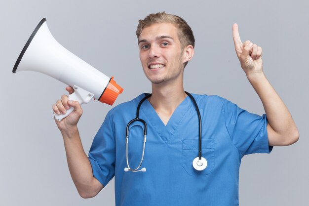 Впечатленный молодой мужчина-врач в униформе врача со стетоскопом, держащим громкоговоритель, смотрит вверх, изолирован на белой стене