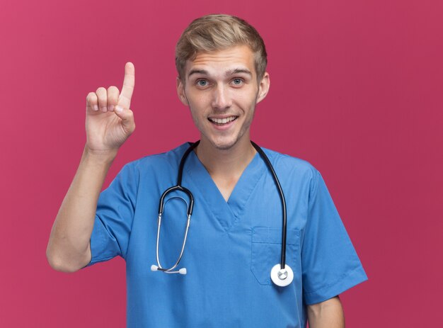 Впечатленный молодой мужчина-врач в униформе врача со стетоскопом указывает вверх на розовой стене
