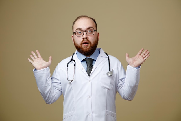 Impressed young male doctor wearing glasses lab coat and stethoscope around his neck looking at camera showing empty hands isolated on olive green background