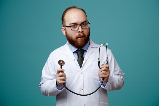 impressed young male doctor wearing medical coat and glasses showing stethoscope looking at camera isolated on blue background