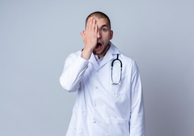 Impressed young male doctor wearing medical robe and stethoscope around his neck covering half of face with hand isolated on white