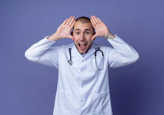 Impressed young male doctor wearing medical robe and stethoscope around his neck keeping hands near head isolated on purple